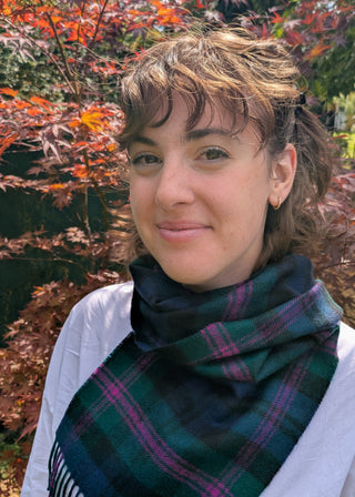 Zu sehen ist ein Klassischer Lammwollschal im Tartan Baird. Dunkelblau, dunkelgrün, schwarz kariert mit Akzenten in Violett. Getragen von einem weiblichen Model in einem Garten. 