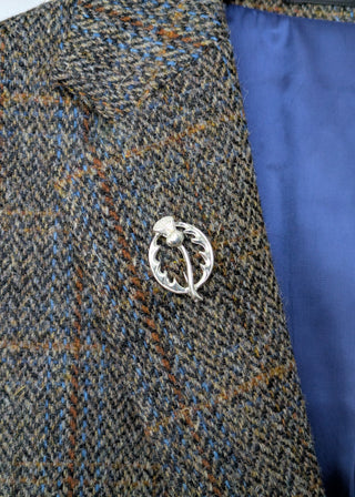 Zu sehen ist eine kleine silberfarbene Anstecknadel in Form einer Distel. Nahaufnahme am Revers eines Harris Tweed Sakkos. 